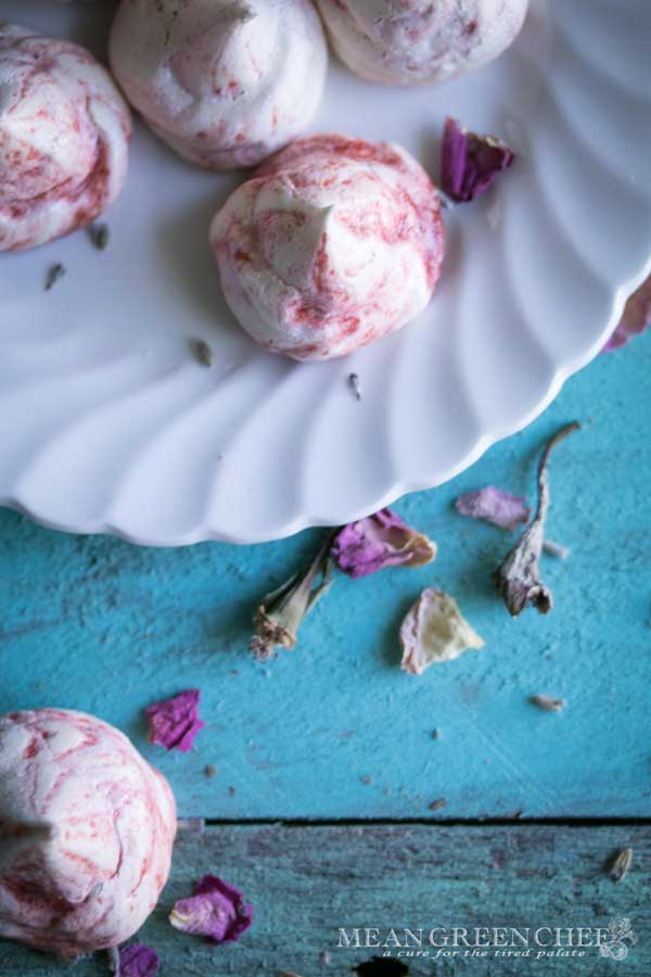 Overhead photo of Strawberry Meringue Cookies sitting on a white plate that's placed on old blue boards and sprinkled with flower petals.