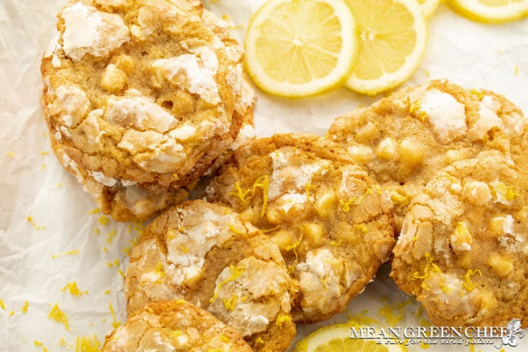 Bright close-up of homemade Meyer Lemon Crinkle Cookies with powdered sugar and lemon zest on top, arranged beside fresh lemon slices on a white surface.