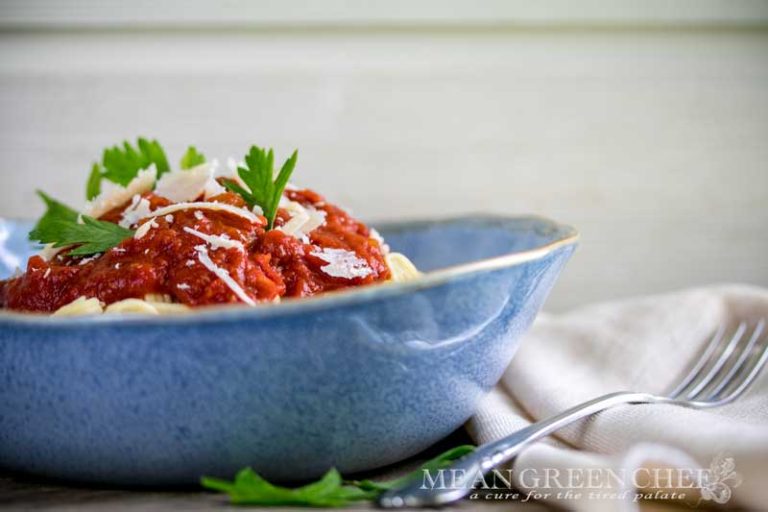 Side photo of a blue bowl piled high with spaghetti and then topped with red auce and garnished with flat leaf parsley and Parmesan.