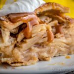 Side photo of a huge slice of Caramel Apple Pie on a white plate garnished with caramel and soft whipped cream. With a pie in the far background. Mean Green Chef