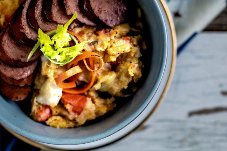 Overhead photo of a bowl of Dutch Stamppot with smoked sausage garnished with carrots and celery sprouts. Mean Green Chef