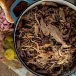 Overhead photo of Slow Roasted Pulled Pork in a gray Dutch Oven with sandwich off the the side in the background. Mean Green Chef