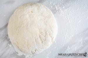 Process photo for making Soft Pretzels, pretzel dough in a disk, sprinkled with flour, on a white marble countertop. Ready to be rolled out.