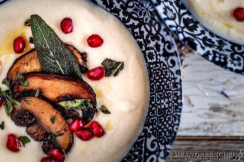 Creamy Cauliflower Bisque ladled into two blue bowls and garnished with smoked portobello mushrooms, fried sage, and pomegranate seeds.