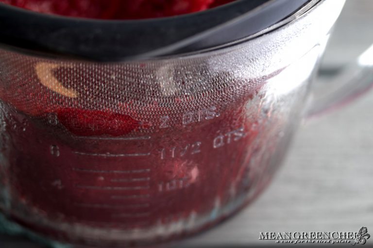 Side photo of raspberries being pushed through a fine mesh strainer into a glass measuring cup.