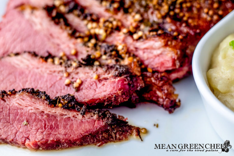 Close-up of a succulent, Guinness-Braised Corned Beef with a peppercorn crust, sliced and displayed on a white plate. With a glimpse of Irish Colcannon to the right-hand side.