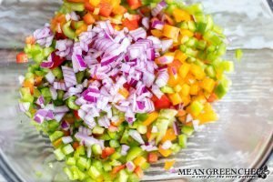 Green, red, and yellow bell peppers being tossed with a diced red onion for chicken pasta salad.