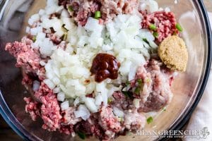 A bowl of raw minced meat mixed with chopped onions, a dollop of Sriracha sauce, and a spoonful of mustard, ready for cooking Larb for Thai Lettuce Wraps.