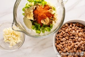 A glass measuring cup of chopped green peppers and spices on a marble countertop, next to a bowl of pinto beans and a small dish of chopped garlic.