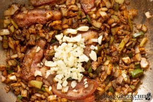 A close-up image of a skillet with chopped onions and pork jowl cooking, with chunks of garlic added on top, showcasing a sizzling, savory meal in preparation.