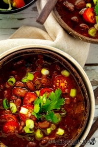 Two bowls of Slow Cooker Texas Pinto Beans garnished with scallions and cilantro, accompanied by a smaller bowl of additional fresh chopped toppings, all presented on a white wooden surface.