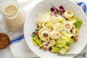 A fresh Big Easy Italian Salad topped with shredded Parmesan cheese, served in an ornate white bowl, accompanied by Creamy Italian Dressing on a white and blue striped cloth.