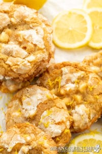 Bright close-up of homemade Meyer Lemon Crinkle Cookies with powdered sugar and lemon zest on top, arranged beside fresh lemon slices on a white surface.