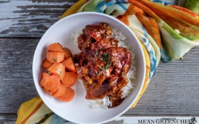 A bowl of Chicken Pierre coated in a rich tomato sauce, served over Jasmine rice. Garnished with fresh thyme, accompanied by a side of roasted carrots. Situated on a rustic wood background, with colorful silk woven underneath the bowl and out of the top of the frame.